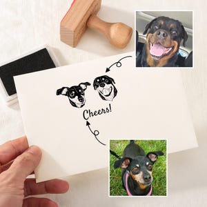 May include: A white card with a black illustration of two dogs' heads and the word "Cheers!" printed on it. The card is being held by a hand. There are two photos of dogs in the background.
