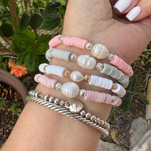 May include: A collection of bracelets in shades of pink, white, and gray. The bracelets feature disc-shaped beads, pearls, and silver accents. One bracelet is a twisted silver bangle, and another has small silver beads.