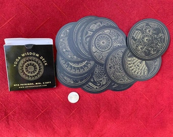 Op de afbeelding: Een zwarte doos met gouden letters die "Yoga Wisdom Deck" en de naam van de maker, "Nta Patrinos, MFA, C-IAYT", lezen. De doos is open en toont een deck ronde zwarte kaarten met gouden ontwerpen. De kaarten zijn in een cirkel gerangschikt.