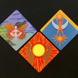 Op de afbeelding: Drie ruitvormige kunstdrukken. E&eacute;n toont een vrouw met vlindervleugels, een andere een vrouw met zon- en maanbeelden, en de derde toont handen die een zon vasthouden. De prints zijn kleurrijk.