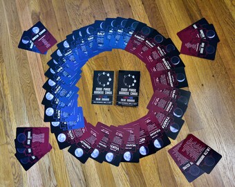 May include: A collection of Moon Phase Goddess Cards arranged in a circular pattern on a wooden surface. The cards are in shades of blue and burgundy, with moon phase illustrations and text. Two decks of cards are in the center, with the text "MOON PHASE GODDESS CARDS".