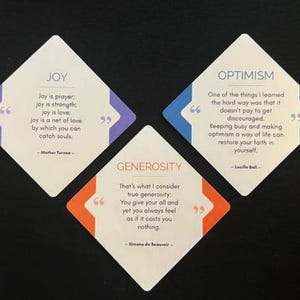 Op de afbeelding: Drie witte ruitvormige kaarten met inspirerende citaten. De kaarten zijn gelabeld met "JOY", "OPTIMISME" en "GENEROSITEIT" met citaten van Moeder Teresa, Lucille Ball en Simone de Beauvoir. Elke kaart heeft een gekleurde rand.