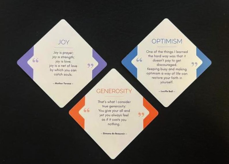 Op de afbeelding: Drie witte ruitvormige kaarten met inspirerende citaten. De kaarten zijn gelabeld met "JOY", "OPTIMISME" en "GENEROSITEIT" met citaten van Moeder Teresa, Lucille Ball en Simone de Beauvoir. Elke kaart heeft een gekleurde rand.