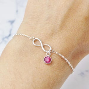 May include: A silver bracelet with an infinity symbol pendant and a small, round, pink gemstone. The bracelet is made of a fine chain. The gemstone is a bright pink colour and is suspended from the infinity symbol.