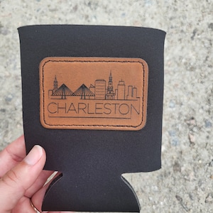 May include: Black neoprene can cooler with a brown leather patch featuring a skyline of Charleston, South Carolina. The patch has the text "CHARLESTON" in black.