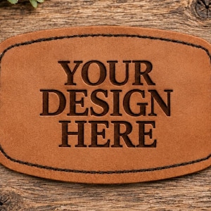 May include: A rectangular tan leather patch with rounded corners and dark brown stitched edges. The center of the patch features the embossed text "YOUR DESIGN HERE" in a bold, capitalized, serif font, creating a clear, recessed appearance.