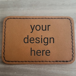 May include: A brown leather patch with black stitching around the edges. The text "your design here" is printed in black on the patch.