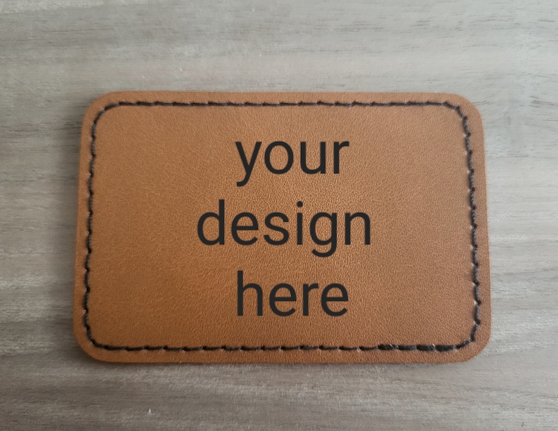 K&ouml;nnte beinhalten: Ein braunes Lederst&uuml;ck mit schwarzen N&auml;hten an den R&auml;ndern. Der Text "your design here" ist in Schwarz auf dem St&uuml;ck aufgedruckt.