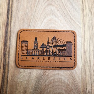 K&ouml;nnte beinhalten: Ein braunes Leder-Patch mit schwarzem Rand und einer schwarzen Silhouette der Skyline von Charleston, South Carolina. Der Text "CHARLESTON" und "SOUTH CAROLINA" ist ebenfalls in Schwarz gedruckt.