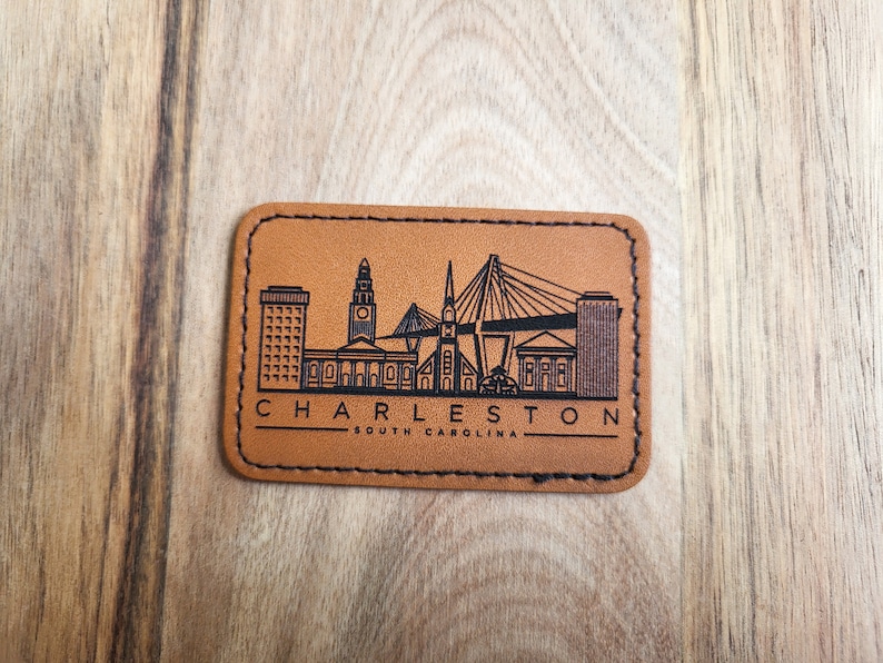 K&ouml;nnte beinhalten: Ein braunes Leder-Patch mit schwarzem Rand und einer schwarzen Silhouette der Skyline von Charleston, South Carolina. Der Text "CHARLESTON" und "SOUTH CAROLINA" ist ebenfalls in Schwarz gedruckt.