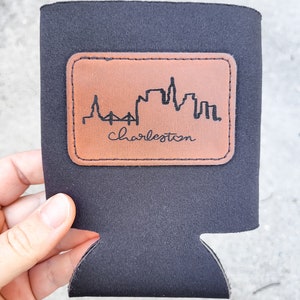 May include: Black neoprene can cooler with a brown leather patch featuring a black silhouette of the Charleston skyline and the text "Charleston".