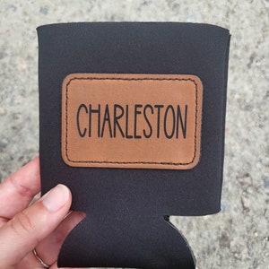 May include: Black neoprene can cooler with a brown leather patch that says "CHARLESTON" in black lettering.