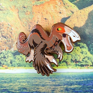 May include: A metal enamel pin shaped like a feathered dinosaur with brown, tan, and cream plumage. The creature has a bright orange patch around its yellow eye, a wide-open mouth with visible teeth, and sharp claws on its feet and wings.