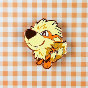 Puede incluir: Un pin con una criatura de dibujos animados naranja y amarilla, con una expresión amigable. El pin tiene un acabado metálico brillante y está sobre un fondo a cuadros naranja y blanco.