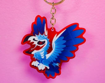 Puede incluir: Un llavero de acrílico rojo y azul con un pájaro estilizado con las alas extendidas. El pájaro tiene una cara blanca con ojos negros y un pico rojo.