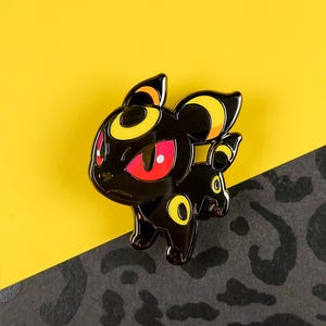 May include: A black enamel pin featuring a stylized cartoon creature with yellow and orange accents. The creature has red eyes and a determined expression. The pin is set against a yellow and black patterned background.