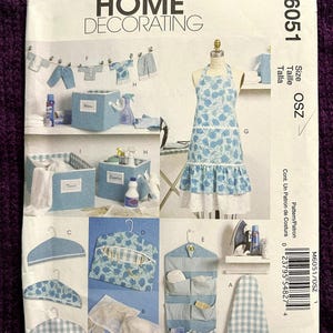 May include: McCall's Home Decorating sewing pattern M6051. The pattern features various home decor items, including an apron, storage boxes, hangers, and organizers. The apron has a blue floral design with a white ruffle. The pattern includes multiple sizes.