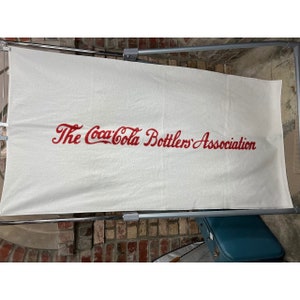 May include: White towel with red embroidered text that reads "The Coca-Cola Bottlers' Association."