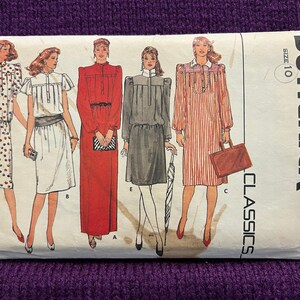 Puede incluir: Un patrón de costura vintage de Butterick Classics, talla 10, con ilustraciones de cinco diseños de vestidos diferentes. Los vestidos varían en estilo, con detalles como lunares, rayas y colores lisos. El patrón está sobre un fondo de punto morado.