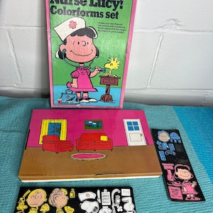 May include: A vintage "Carry on, Nurse Lucy! Colorforms set" featuring Peanuts characters. The box shows Lucy in a nurse's uniform with Woodstock. Includes a room scene and character cutouts for creative play. The set is in a pink and green box.