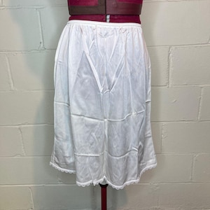 May include: A white slip skirt with a scalloped lace hem is displayed on a maroon dress form. The skirt is made of a lightweight, slightly sheer fabric. The waistline is elasticized and has a delicate trim.