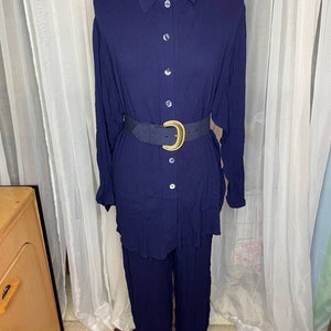 Op de afbeelding: Een donkerblauwe blouse met knopen en een bijpassende broek. De blouse heeft lange mouwen en een kraag. De broek heeft wijde pijpen en een ontspannen pasvorm. Een bruine leren riem met een gouden gesp wordt om de taille gedragen.