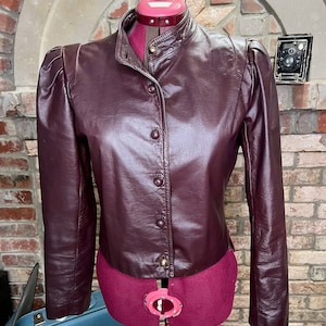 May include: A maroon leather jacket with a stand-up collar, long sleeves, and a button-up front. The jacket has a cropped fit and a slightly puffed shoulder design.