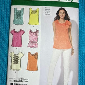 Peut inclure: Patron de couture Simplicity 2599. Le patron comprend des modèles de hauts avec différents cols et longueurs de manches. La taille du patron est D5 4,6,8,10,12, et EUR 30-38, FR 32-40. L'image montre une femme portant un haut pêche et un pantalon blanc.