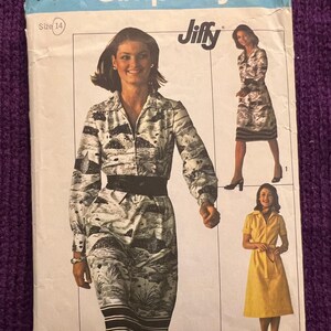 Puede incluir: Un sobre de patrón de costura Simplicity vintage, talla 14, con una mujer modelando un vestido con estampado de paisaje en blanco y negro. El sobre incluye imágenes de otros estilos de vestidos y el texto "Jiffy". El fondo es de tejido de punto morado.