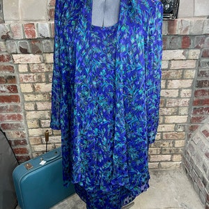 May include: A vintage blue dress and jacket set on a mannequin. The dress and jacket feature a floral pattern in shades of blue and teal. The long-sleeved jacket is draped over the dress, creating a layered look. The dress is knee-length.