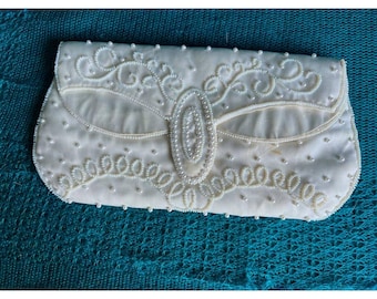Könnte beinhalten: Eine weiße Clutch aus Satin mit einem Perlmutt-Perlenmuster. Die Clutch hat einen Klappverschluss und eine perlenbesetzte ovale Form in der Mitte.