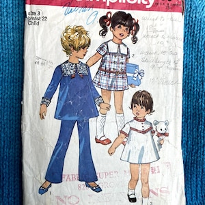 May include: Simplicity sewing pattern number 8567 for children's clothing. The pattern includes three different styles: a blue dress with a white collar, a plaid dress with a white collar, and a white dress with a red collar. The pattern is for sizes 16 to 20 inches.