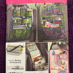 May include: Simplicity pattern 2916 for car seat organizers. The pattern features various designs, including organizers with multiple pockets, a purse, and a baby organizer. The organizers have colorful patterns and are designed by Robin Greenwood.