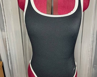 Puede incluir: Traje de baño negro con ribete blanco alrededor del escote y las aberturas de las piernas. El traje de baño está en un maniquí rojo.