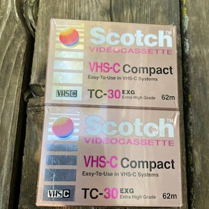 Könnte beinhalten: Zwei VHS-C-Videokassetten von Scotch. Die Kassetten sind goldfarben und rosa mit dem aufgedruckten Text "Scotch VIDEOCASSETTE VHS-C Compact Easy-To-Use in VHS-C Systems TC-30 EXG Extra High Grade 62m".