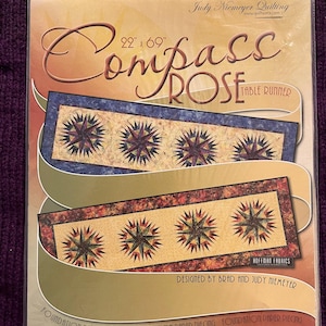 Op de afbeelding: Een pakket voor een Compass Rose tafelloper quiltpatroon. Het ontwerp heeft een sterrenpatroon in tinten blauw, rood en goud. De tafelloper meet 56 cm x 175 cm. Ontworpen door Brad en Judy Niemeyer.