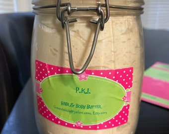 Op de afbeelding: Een glazen pot met een metalen sluiting gevuld met een lichtbruine, romige substantie. De pot heeft een roze en groen etiket met de tekst "P.K.I. Hair & Body Butter. Greenatalicia@yahoo.com, Etsy.com".