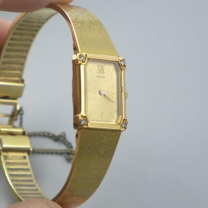 Puede incluir: Un reloj de pulsera Seiko dorado con una esfera rectangular y una correa a juego. La esfera del reloj presenta números romanos y está adornada con pequeños cristales en cada esquina. La correa tiene un diseño texturizado y flexible.