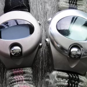 May include: Two silver-toned wristwatches with digital displays and black bands. The watches have a round face with a large, oval-shaped window for the display.