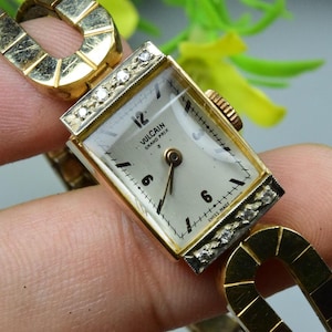 Puede incluir: Reloj de pulsera vintage Vulcain Grand Prix dorado con esfera rectangular y detalles de diamantes. La esfera blanca tiene números y agujas negras. La correa del reloj también es dorada.