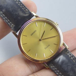 Pode incluir: Um relógio Seiko Dolce dourado com uma pulseira de couro preta. O mostrador do relógio é dourado com ponteiros e marcadores pretos. A pulseira é de couro preto com costuras brancas.