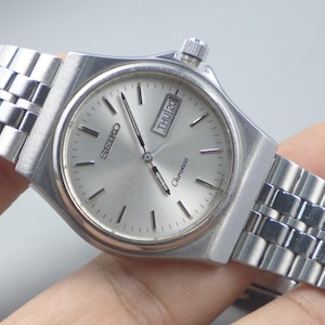 Op de afbeelding: Een zilveren Seiko Chronos polshorloge met een zilveren wijzerplaat en een roestvrijstalen band. De wijzerplaat toont de dag en datum. Het horloge heeft een rechthoekige kast en een getextureerde kroon.