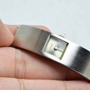 May include: A silver-toned cuff watch with a wide, brushed metal band and a small, square watch face. The dial is light-colored with the word GUESS printed in black, featuring two thin hands and a small winding crown on the side of the casing.