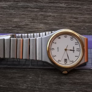 Könnte beinhalten: Eine silberne und goldene Seiko Spirit Uhr mit weißem Zifferblatt und schwarzen Zeigern. Die Uhr hat ein Datumsfenster und ein silbernes Metallband.