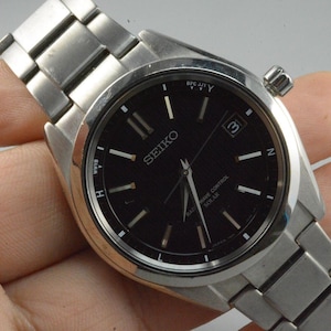 Könnte beinhalten: Eine Seiko-Uhr aus Edelstahl mit schwarzem Zifferblatt. Die Uhr hat silberne Zeiger und Markierungen, ein Datumsfenster bei 3 Uhr und ein silbernes Armband. Die Uhr wird von einer Hand gehalten.
