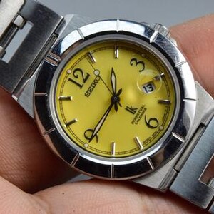 May include: A Seiko wristwatch with a bright yellow dial, silver-tone hands, and black Arabic numerals at the 12, 3, 6, and 9 o'clock positions. The watch features a stainless steel case and link bracelet, a date window, and the text Perpetual Calendar.