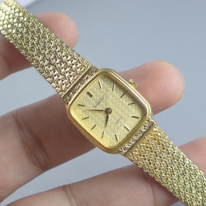 Könnte beinhalten: Goldfarbene Aureole-Quarzuhr mit rechteckigem Zifferblatt und strukturiertem Zifferblatt. Die Uhr hat ein goldenes Mesh-Armband. Die Zeiger und Stundenmarkierungen sind schwarz. Ein klassisches Accessoire.