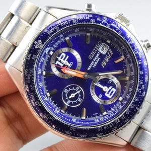 May include: A silver-tone stainless steel Seiko chronograph watch with a vibrant blue dial. The watch features a rotating slide rule bezel, three sub-dials, a date window at the three o'clock position, and luminous hands with an orange-tipped second hand.