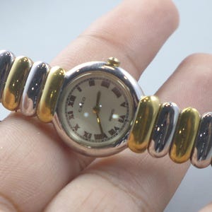 Könnte beinhalten: Eine silber- und goldfarbene Quarzuhr mit cremefarbenem Zifferblatt und römischen Ziffern. Das Uhrenarmband ist im Stretch-Stil gehalten und wechselt zwischen silbernen und goldfarbenen Gliedern. Die Uhr wird in einer Hand gehalten.