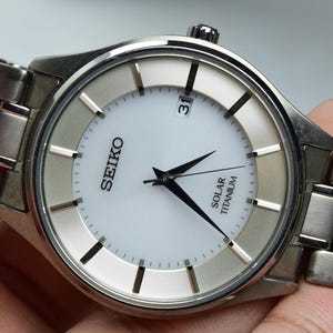 May include: A Seiko Solar Titanium wristwatch featuring a round white dial with black hour markers and hands. The watch includes a date window at the three o'clock position and a silver-toned metal link bracelet. The text Seiko and Solar Titanium are printed on the face.
